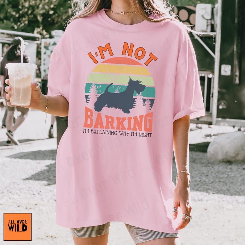 May include: A light pink t-shirt featuring a graphic of a black Scottish Terrier silhouette centered within a retro-style sunset circle. The text reads I'M NOT BARKING I'M EXPLAINING WHY I'M RIGHT in orange and black lettering with a forest background.