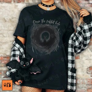 May include: Black t-shirt featuring the text "Down the rabbit hole" and a dark circular design. A black and white checked cardigan is worn over the shirt. A plush black bat with pink ears is held in front of the shirt.