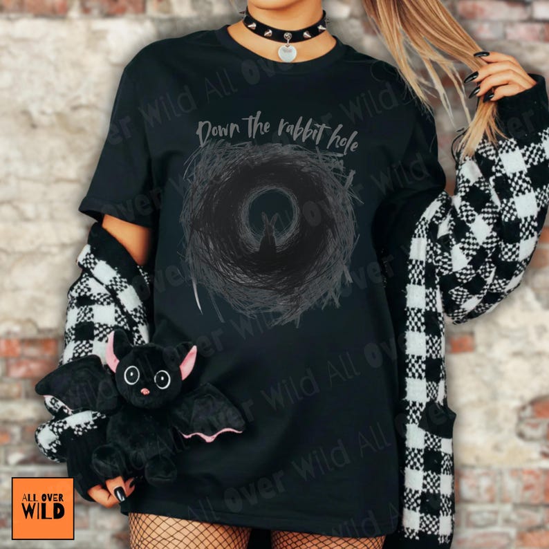 May include: Black t-shirt featuring the text "Down the rabbit hole" and a dark circular design. A black and white checked cardigan is worn over the shirt. A plush black bat with pink ears is held in front of the shirt.