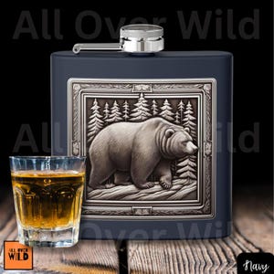 May include: Navy blue flask with a silver cap and a detailed bear and forest design. A shot glass with amber liquid sits in front of the flask. The words "All Over Wild" are visible.