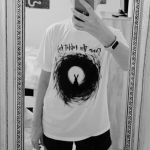 May include: White t-shirt featuring a black rabbit silhouette within a circular design, with the text "Down the Rabbit Hole" above. The shirt is worn with black shorts. The image is captured in front of a mirror.