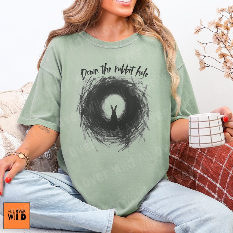 May include: Sage green t-shirt with a black graphic of a rabbit in a dark circular design, and the text "Down the rabbit hole" above. The shirt has a relaxed fit and short sleeves. The brand logo "ALL OVER WILD" is visible.