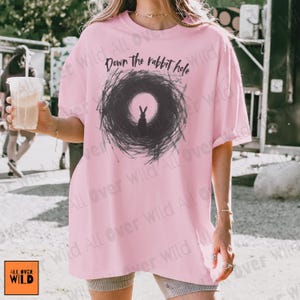 May include: A pale pink t-shirt featuring the phrase "Down the rabbit hole" above a black and grey illustration of a rabbit. The shirt has a loose fit and short sleeves. The brand logo "ALL OVER WILD" is visible in the bottom left corner.