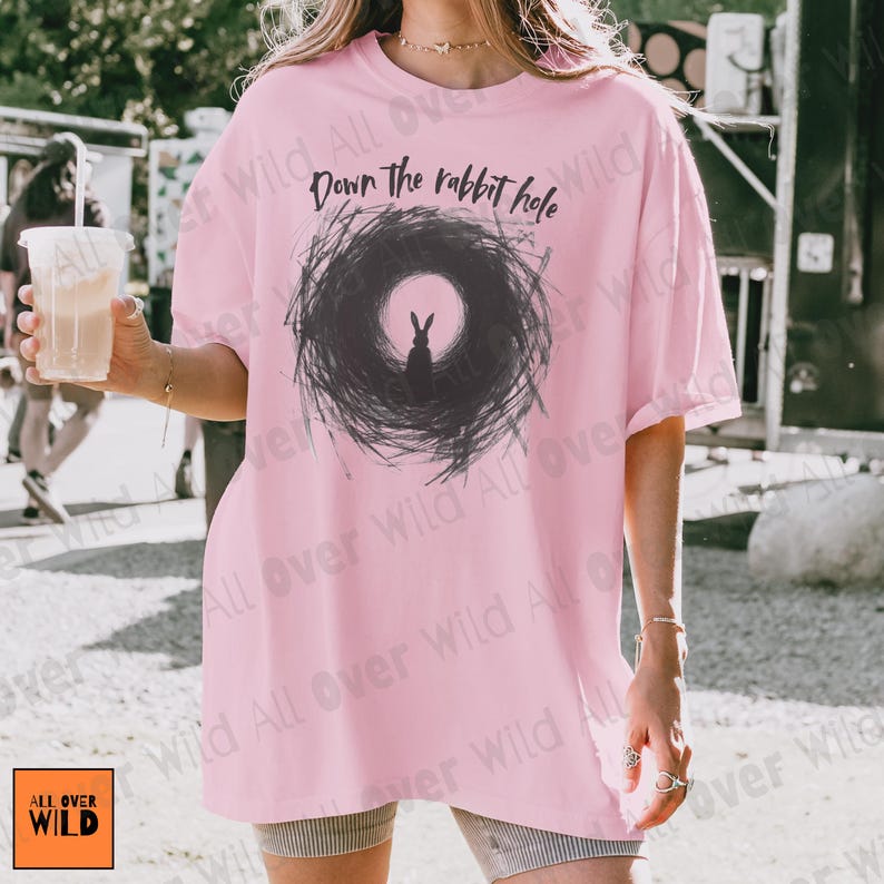 May include: A pale pink t-shirt featuring the phrase "Down the rabbit hole" above a black and grey illustration of a rabbit. The shirt has a loose fit and short sleeves. The brand logo "ALL OVER WILD" is visible in the bottom left corner.