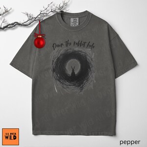 May include: Grey t-shirt featuring the phrase "Down the rabbit hole" and a black rabbit graphic within a swirling design. The tee has a vintage wash and a loose fit. The brand logo "All Over Wild" is visible in the bottom left corner.