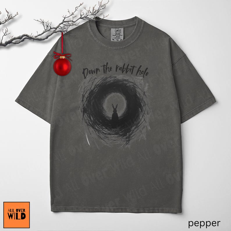 May include: Grey t-shirt featuring the phrase "Down the rabbit hole" and a black rabbit graphic within a swirling design. The tee has a vintage wash and a loose fit. The brand logo "All Over Wild" is visible in the bottom left corner.