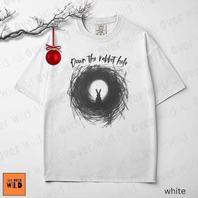 May include: White t-shirt featuring the phrase "Down the rabbit hole" above a black and grey rabbit graphic. The tee has a crew neck and short sleeves. The brand logo "ALL OVER WILD" is in the bottom left corner.
