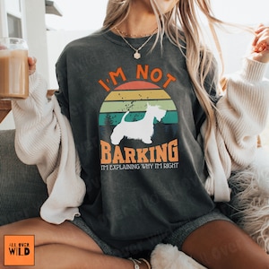 May include: A charcoal gray crewneck sweatshirt featuring a graphic of a white Scottish Terrier silhouette centered within a retro-style sunset circle. The text reads I'M NOT BARKING I'M EXPLAINING WHY I'M RIGHT in orange and white lettering.