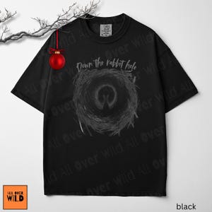 May include: Black t-shirt featuring the text "Down the rabbit hole" above a grey spiral design. The shirt has a crew neck and short sleeves. A red Christmas bauble hangs from a branch in the upper left.