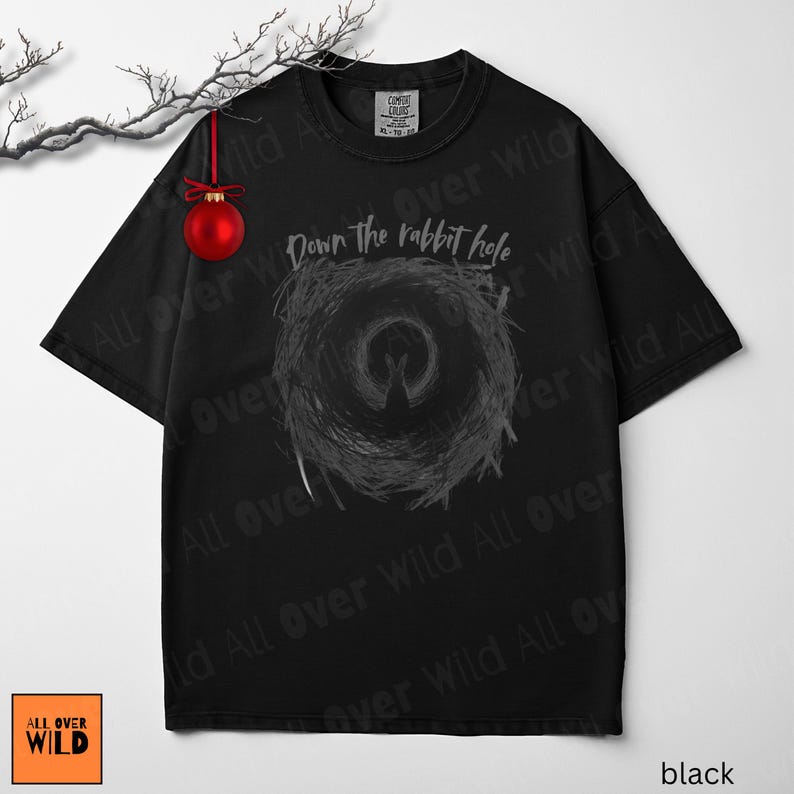 May include: Black t-shirt featuring the text "Down the rabbit hole" above a grey spiral design. The shirt has a crew neck and short sleeves. A red Christmas bauble hangs from a branch in the upper left.