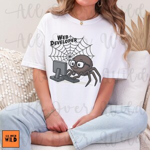 Può includere: T-shirt bianca con un ragno dei cartoni animati con gli occhiali che lavora al computer. Il ragno è di fronte a una ragnatela con le parole "WEB DEVELOPER" sopra. La maglietta ha il logo "ALL OVER WILD".
