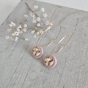 Puede incluir: Un par de pendientes de aro dorados con un colgante circular de gamuza rosa en cada aro.