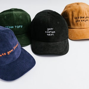 May include: Four corduroy baseball caps in different colors with embroidered text. The caps are green, black, blue, and brown. The text on the caps reads "Team Tofu", "Your custom text", "Hats your strain!" and "Coffee yes you maybe."
