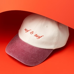 Può includere: Un cappellino da baseball bianco con visiera bordeaux. La scritta "surf & turf" è ricamata in rosso sul davanti. Il cappellino è posto su uno sfondo arancione brillante.