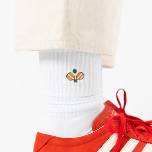 May include: White ribbed socks featuring an embroidered orange and green design, worn with red trainers with white stripes. The socks are visible below beige trousers.