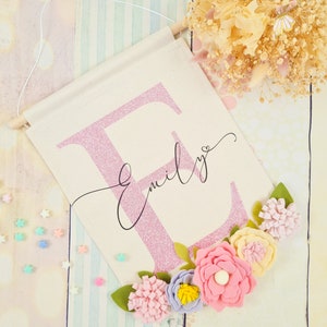 Könnte beinhalten: Ein pinkfarbener Glitzerbuchstabe E mit dem Namen "Emily" darunter in Kursivschrift. Der Buchstabe ist von einem Kranz aus Filzblumen in den Farben Rosa, Gelb und Lila umgeben.