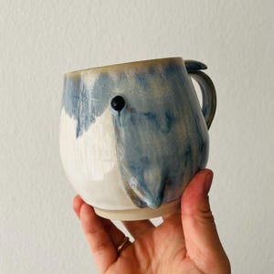 Könnte beinhalten: Ein Keramikbecher mit abgerundetem Körper und einer glänzenden Glasur in Weiß- und Blau-Tönen. Der Becher hat einen Henkel auf der rechten Seite und eine kleine, dunkle, kreisförmige Erhebung auf der oberen Vorderseite, die einem Auge ähnelt.