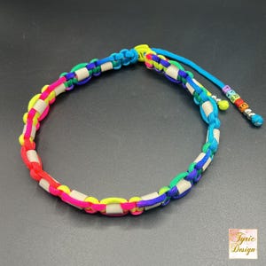 Könnte beinhalten: Ein Regenbogen-Paracord-Armband mit weißen Perlen und einem kleinen Anhänger mit den Buchstaben "TYRIC" darauf.