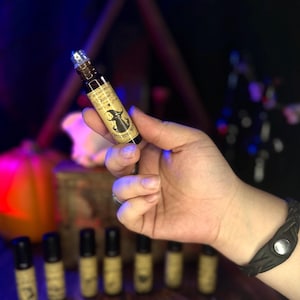 Op de afbeelding: Een verzameling kleine glazen flesjes met roll-on applicators. De flesjes zijn gevuld met een donkere vloeistof en hebben etiketten met een heks en maan ontwerp. De etiketten lezen "Witching Hour" en "Autumn Moon".