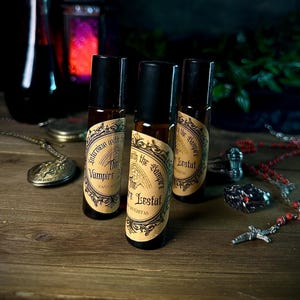 May include: Three brown glass roll-on bottles with black caps and vintage-style labels. The labels read "Interview with the Vampire" and "The Vampire Lestat". The bottles are on a wooden surface with gothic-themed accessories.