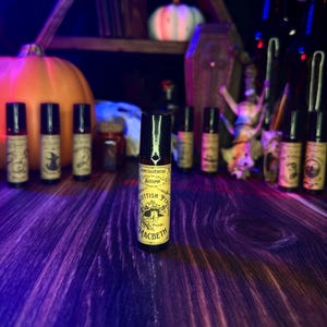Puede incluir: Un frasco negro de roll-on con una etiqueta amarilla que dice "Fantasveritas Autumn Scottish Play Macbeth". El frasco está sobre una superficie de madera con una calabaza y otras botellas en el fondo.