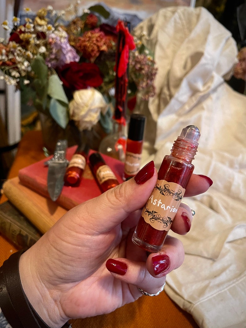 May include: A red glass bottle with a rollerball applicator and a gold label that says "Astarian". The bottle is filled with a red liquid and has glitter inside.