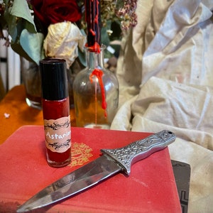 May include: A small glass bottle with a red liquid and a label that says "Astarian" is sitting on a red book. A silver knife with an ornate handle is lying on top of the book.