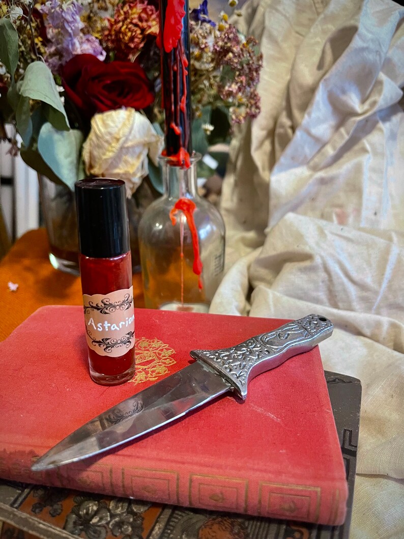May include: A small glass bottle with a red liquid and a label that says "Astarian" is sitting on a red book. A silver knife with an ornate handle is lying on top of the book.