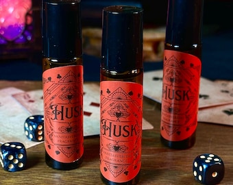 May include: Three glass roll-on bottles with black caps and red labels, each labeled "Husk Vanitasveritas Fragrance." The bottles are on a wooden surface with playing cards and dice. The background includes a bottle of wine.