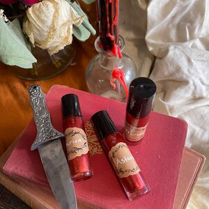May include: Three small glass bottles with red glitter liquid and black roll-on tops. Each bottle has a brown label with a decorative border and white text. The labels read "Astarte", "Marion", and "Marion". The bottles are sitting on a stack of red and brown books. A silver knife is lying on the books.