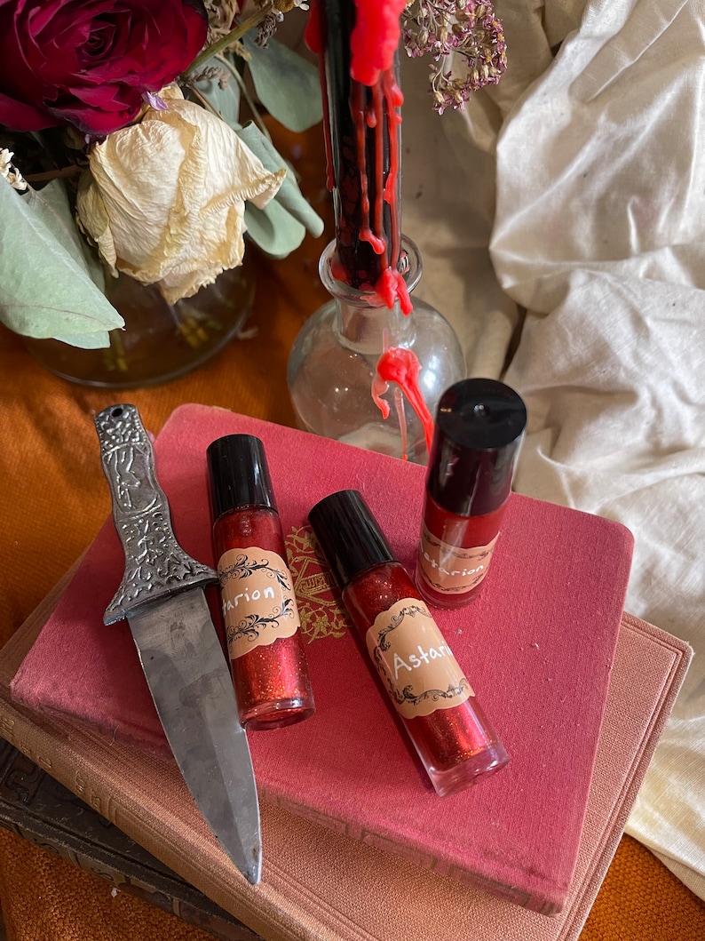 May include: Three small glass bottles with red glitter liquid and black roll-on tops. Each bottle has a brown label with a decorative border and white text. The labels read "Astarte", "Marion", and "Marion". The bottles are sitting on a stack of red and brown books. A silver knife is lying on the books.