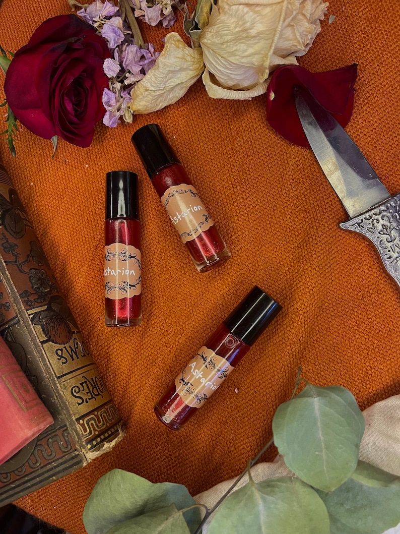 May include: Three small glass bottles with black caps and red liquid inside. Each bottle has a label with the word "Astariel" written in a decorative font. The bottles are on a red surface with dried flowers and a knife.