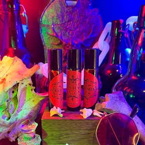 Könnte beinhalten: Drei kleine Glasflaschen mit schwarzen Roll-on-Kappen und roten Etiketten. Die Etiketten tragen weißen Text, der "The Radio Demon", "The Radio Demon" und "The Radio Demon" lautet. Die Flaschen stehen auf einer Holzoberfläche mit einem Schädel und Knochen im Hintergrund.