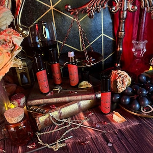 May include: Four small glass bottles with roll-on applicators filled with dark liquid. The bottles have black labels with white text that reads "The Darkest Desire" and a bat graphic. The bottles are arranged on a stack of old books with a gold chain and a nail on a wooden surface.