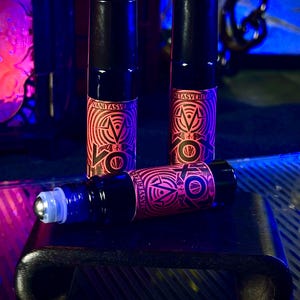 May include: Three black roll-on bottles with red and pink geometric designs and the text "VANITASVERITAS". One bottle is lying down, revealing a silver rollerball. The bottles are on a black surface, with a dark, neon-lit background.