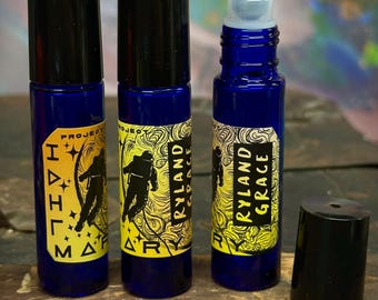 May include: Three cylindrical cobalt blue glass roller bottles with black caps. Each bottle features a yellow label with black text reading Project Hail Mary and Ryland Grace, accompanied by a graphic illustration of an astronaut in space.