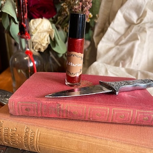 May include: A small, clear glass bottle with a red liquid inside. The bottle has a black cap and a brown label that says "Astarion". The bottle is sitting on top of a stack of three books. A silver knife is lying on top of the books.