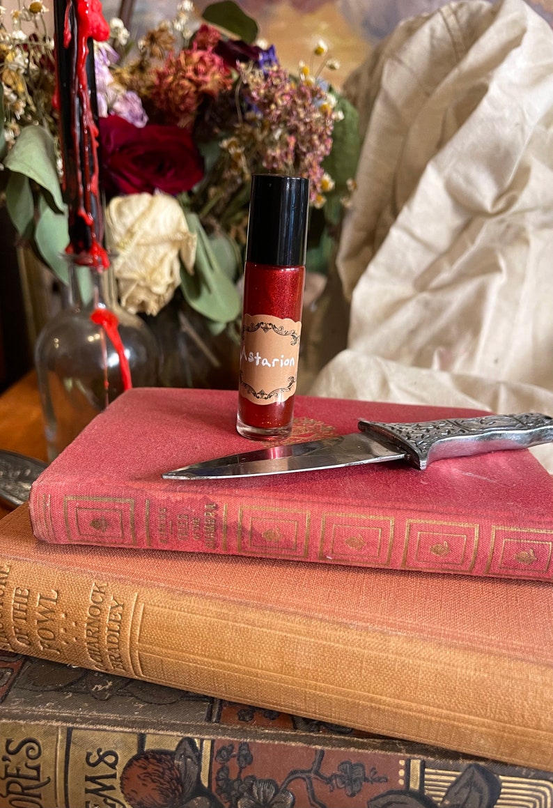 May include: A small, clear glass bottle with a red liquid inside. The bottle has a black cap and a brown label that says "Astarion". The bottle is sitting on top of a stack of three books. A silver knife is lying on top of the books.
