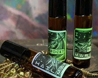 May include: Three amber glass roller bottles with black caps, each featuring a label with the text Project Hail Mary. Two bottles stand upright, while one lies horizontally among a pile of small, metallic gold-colored screws.