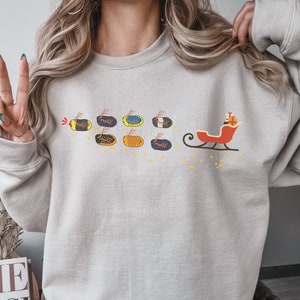 Puede incluir: Sudadera beige claro con un diseño festivo. El gráfico representa un trineo rojo con un regalo, tirado por una serie de calabazas decoradas. Las calabazas son de varios colores y patrones, con estrellas amarillas y un rastro de luz roja.