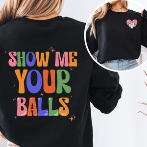 May include: Black sweatshirt with the text "SHOW ME YOUR BALLS" in colorful, retro-style lettering on the back. A smaller image of the sweatshirt is shown on the right, featuring a heart with a cat graphic.