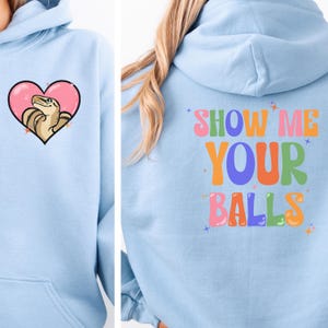 May include: Light blue hoodie with a cartoon snake inside a pink heart on the front. The back of the hoodie has the text "SHOW ME YOUR BALLS" in colorful, retro-style lettering. A casual, fun apparel item.