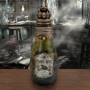 May include: A decorative glass bottle with a green and yellow mottled design. The bottle is labeled "TOADS WART" and has a frog-shaped stopper. The bottle is wrapped with twine and has a vintage apothecary aesthetic.