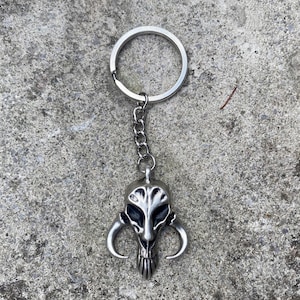 Könnte beinhalten: Ein silberner Schlüsselanhänger mit einem detaillierten Mandalorianer-Schädel-Design. Der Schädel hat ein schwarzes Auge und eine silberne Kette, die an einem silbernen Ring befestigt ist.