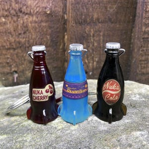 Może przedstawiać: Trzy miniaturowe breloczki w kształcie butelek z Nuka Colą, Nuka Cherry i Quantum. Butelki są czerwone, czarne i niebieskie z białymi etykietami i mają kształt rakiet.