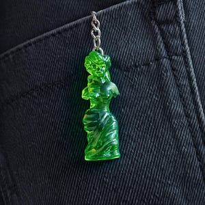 May include: A green, translucent figurine of a classical female bust, hanging from a silver chain. The figure is detailed with flowing drapery and set against a dark denim background. A unique keychain.