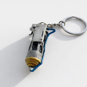 Op de afbeelding: Een gedetailleerde sleutelhanger met een miniatuur, gestileerd apparaat met een zilverkleurig lichaam, gele en blauwe accenten en een zilveren sleutelring. Het ontwerp omvat ingewikkelde details en een kleine ketting die het apparaat met de ring verbindt.
