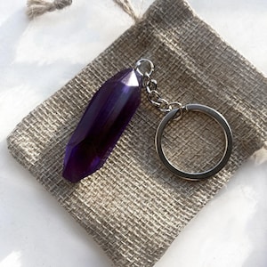 May include: A purple crystal keyring with a silver chain and key ring. The crystal is a long, faceted shape. The keyring rests on a small, tan hessian bag.