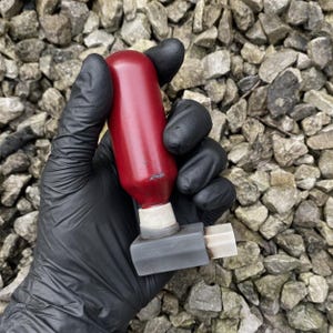 May include: A red bottle stopper with a gray base and a white ring. The stopper is held by a gloved hand against a background of gray rocks. The stopper is cylindrical in shape.