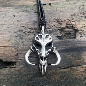 Könnte beinhalten: Eine silberne Halskette mit einem detaillierten Totenkopf mit Hörnern. Der Anhänger hängt an einer schwarzen Lederkordel.